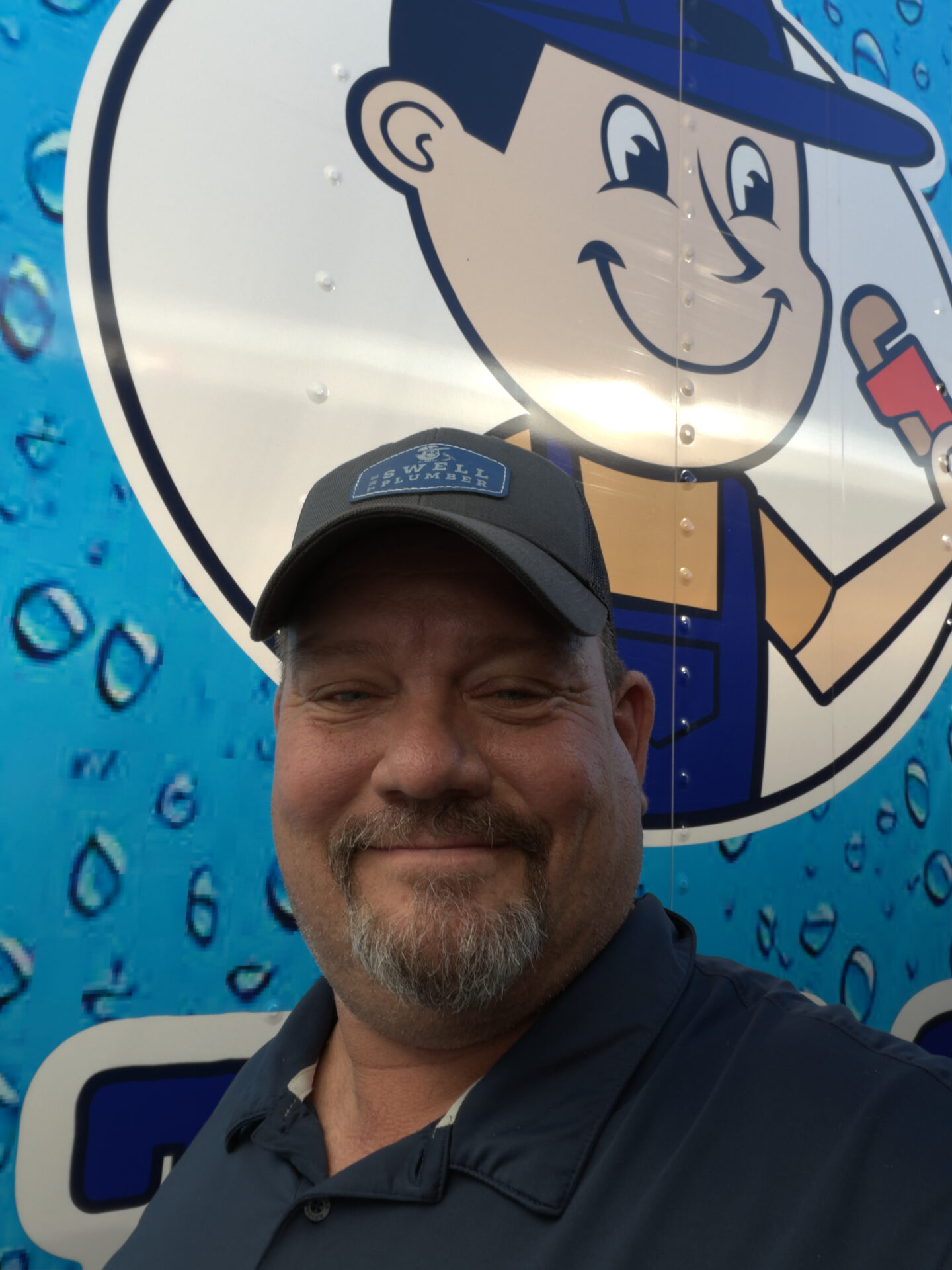 Man in a blue shirt and cap smiling in front of a cartoon handyman graphic on a vehicle with a blue, water-themed background.