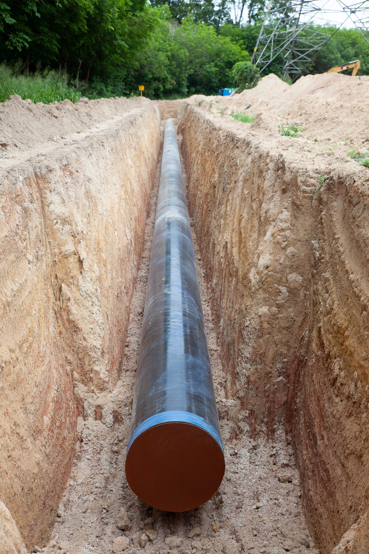 A large metal pipeline is laid in a freshly dug trench in a rural area, surrounded by dirt and vegetation.