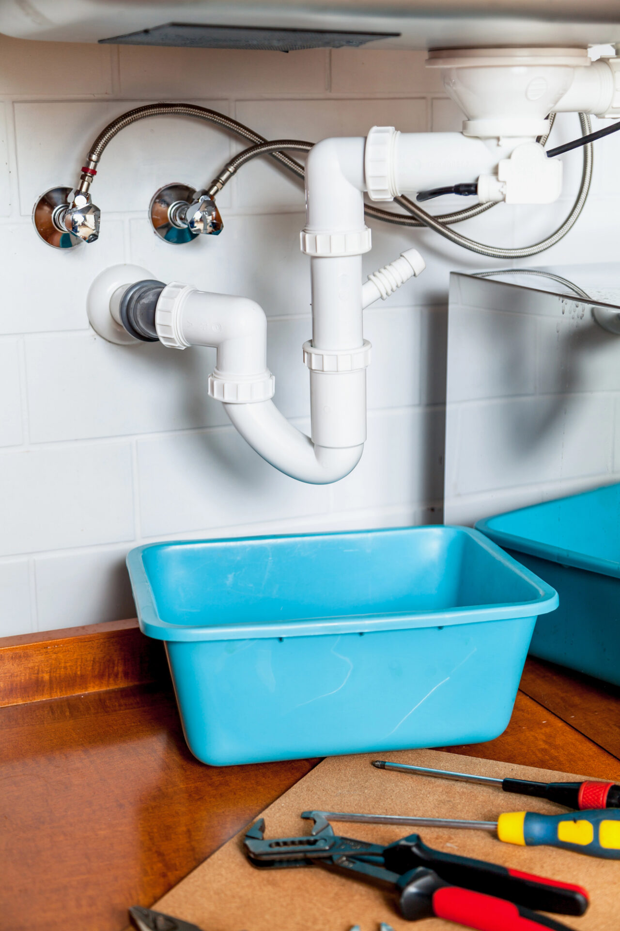 White PVC pipes under a sink with a blue plastic basin below. Plumbing tools and a screwdriver are on the wooden countertop.