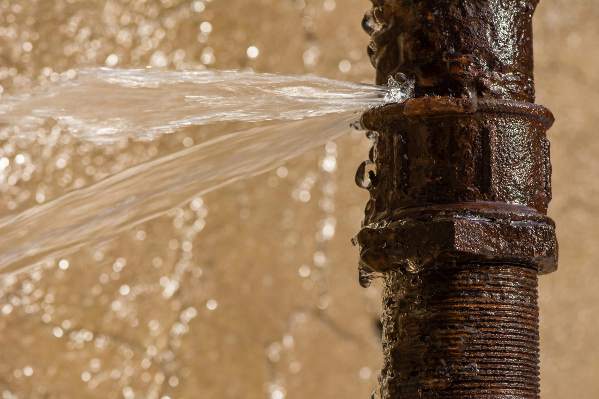 Water sprays out of a crack in a rusty metal pipe against a blurred background.