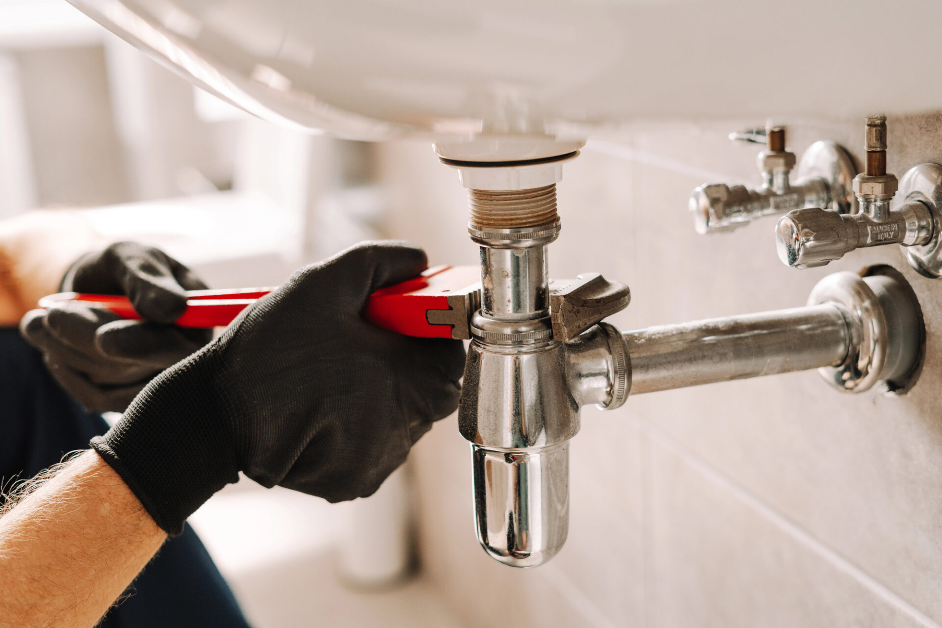 Person wearing black gloves using a red wrench to tighten or loosen a pipe under a bathroom sink with visible plumbing fixtures.