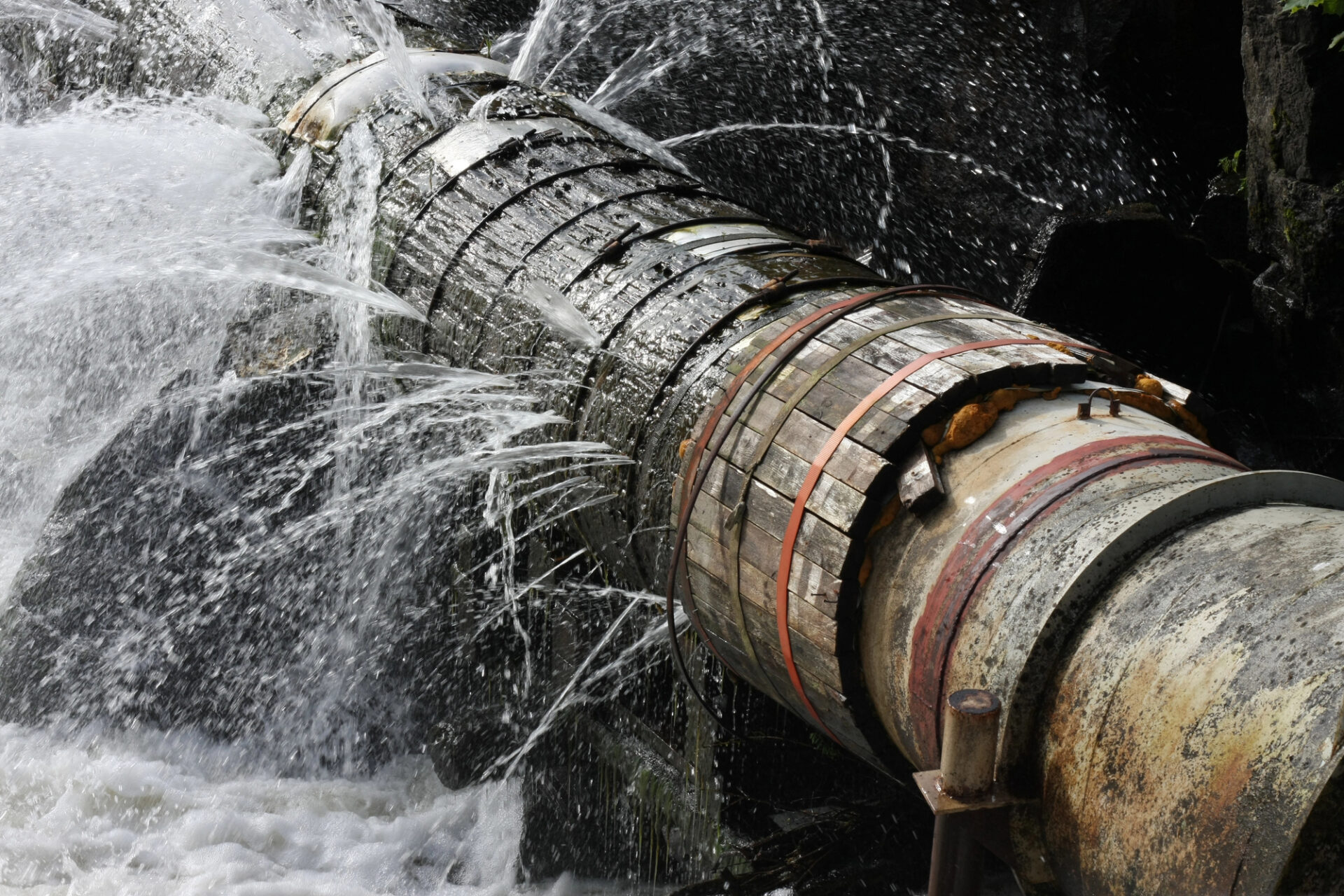 A large metal pipe with multiple leaks sprays water in several directions, indicating serious damage or failure in the structure.