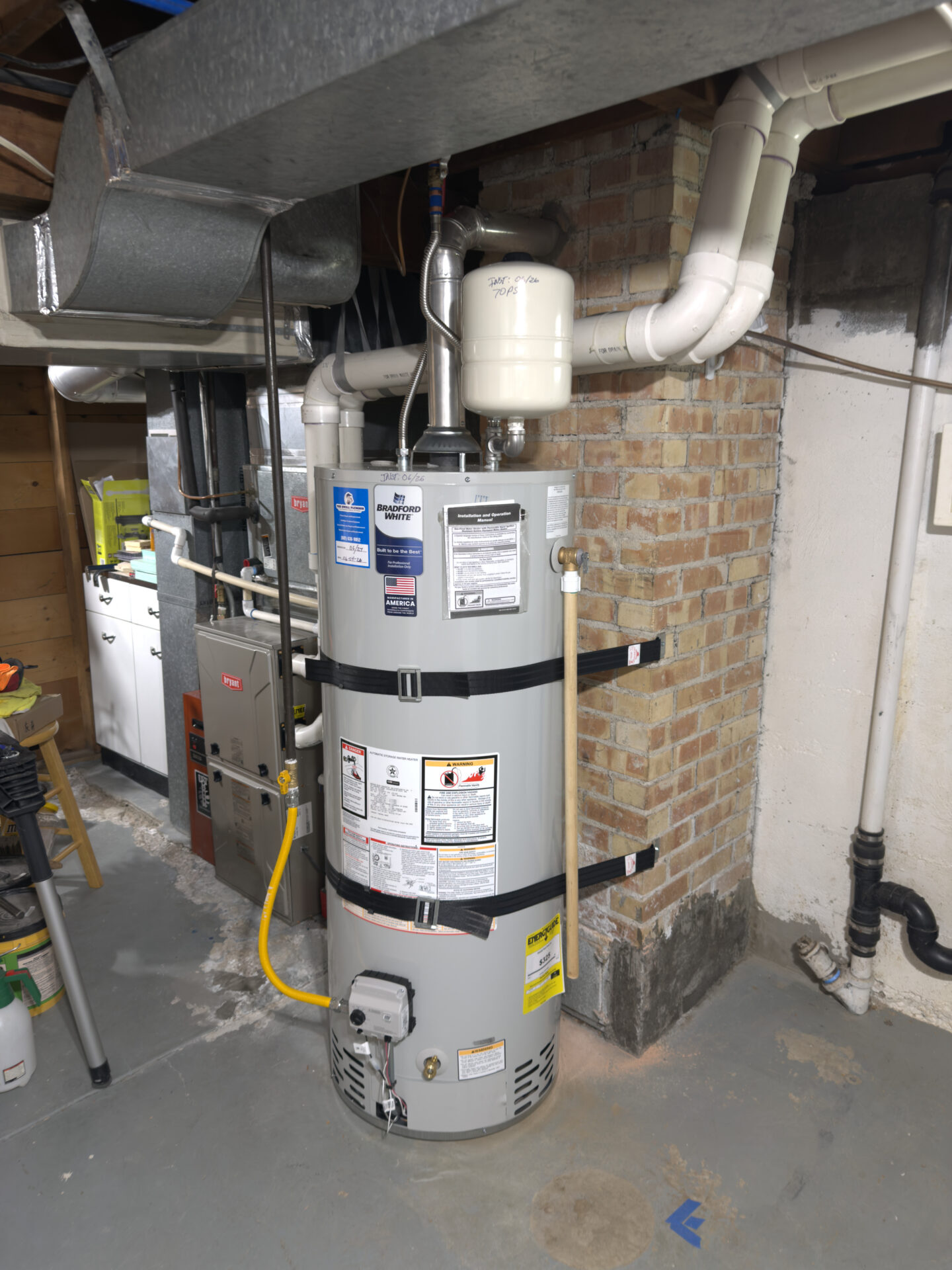 A gas water heater with attached expansion tank is installed in a basement with exposed brick walls and visible ductwork.